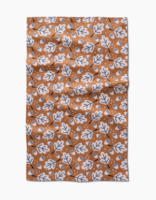 A rectangular light brown Charlie Rowan Designs Falling Acorns tea towel, made from post-consumer recycled materials, features a white oak leaf and small acorn pattern outlined in black and is shown flat with slight wrinkles.