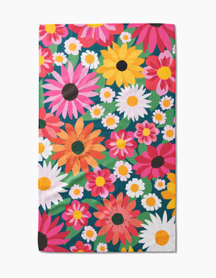 The Wild Blooms Tea Towel by Rebecca Bobko features a vibrant floral pattern in pink, yellow, white, and orange with green leaves—crafted from post-consumer recycled materials for eco-friendly style.