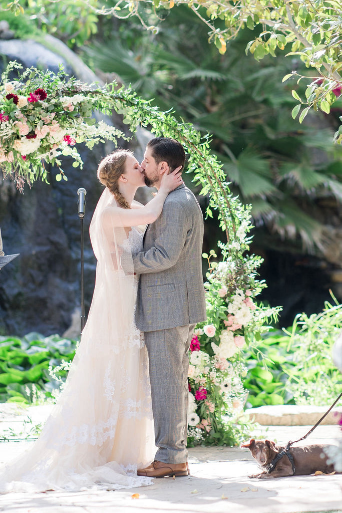 champagne swiss dot veil and blush nude bridal gown on bride kissing groom in front of rose flower arch on wedding day