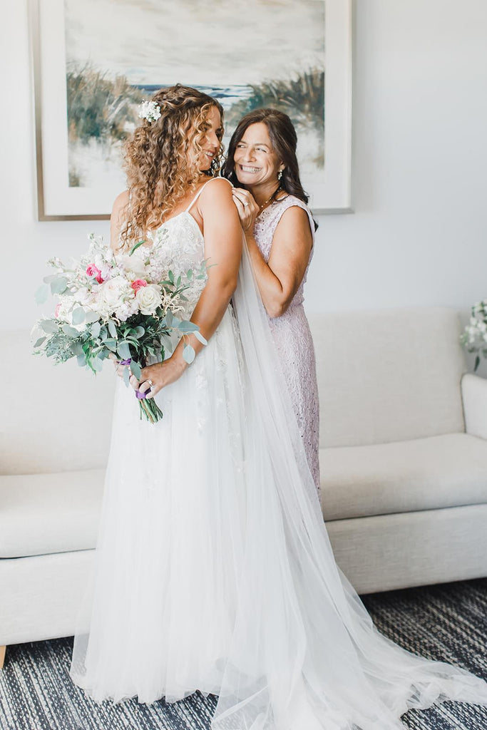 beautiful curly haired bride wearing long off white bridal tulle wing set as mother helps