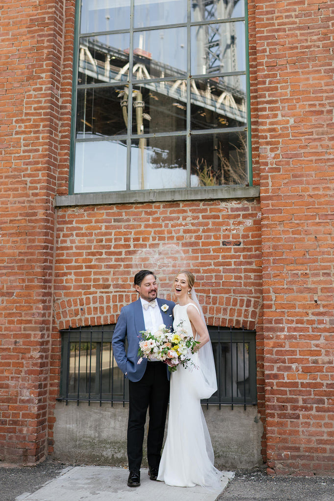 Laughing bride in minimalist simple crepe sheath dress and silk cathedral veil with groom in front of red brick east coast building