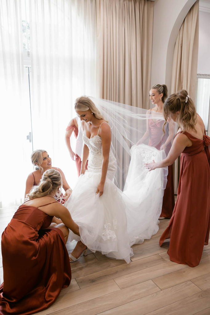 glam bride getting dressed in mermaid gown and fingertip sparkly glimmer veil as bridesmaids surround her in maroon red gowns