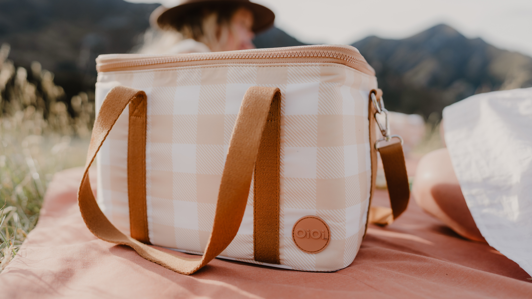a beige gingham cooler bag sits on a pink rug in a family picnic scene