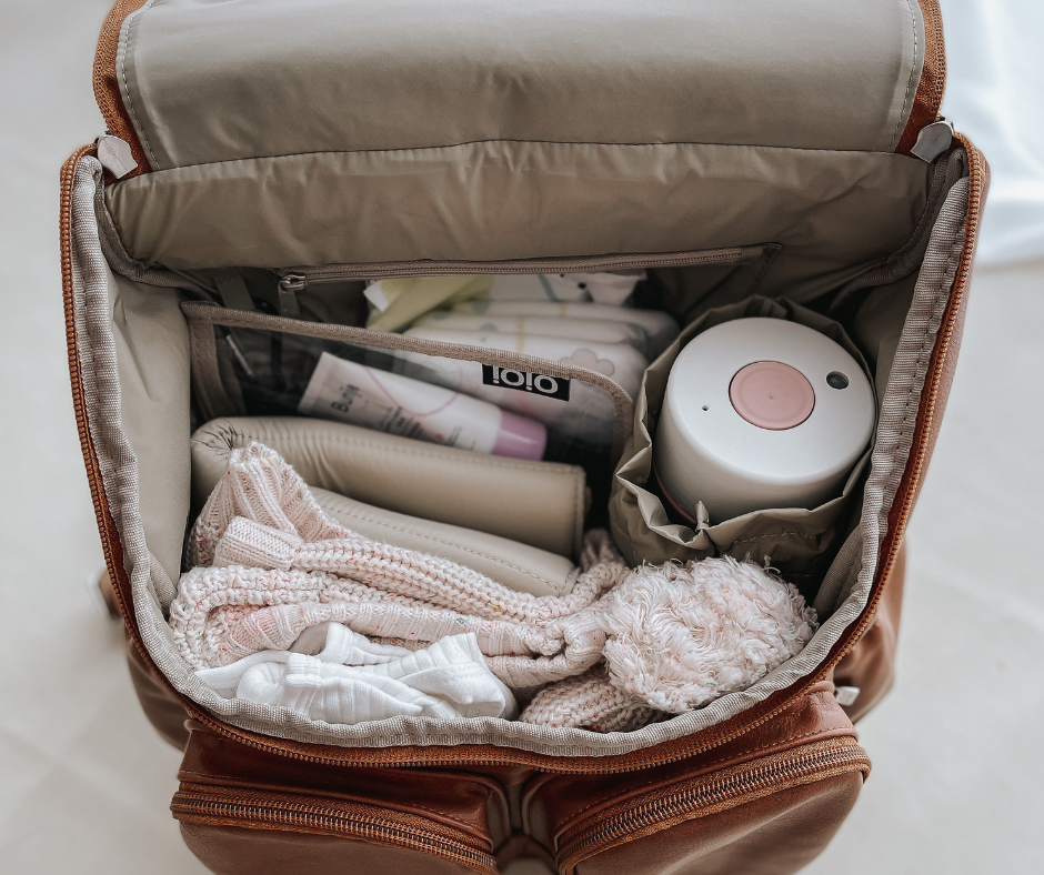 A packed nappy bag for a newborn, containing diapers, wipes, extra clothes, snacks, toys, sunscreen, hats, a breast pump and bottles, and a first aid kit.