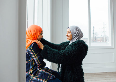 two sisters wearing an orange and grey hijab talking to each other in a home