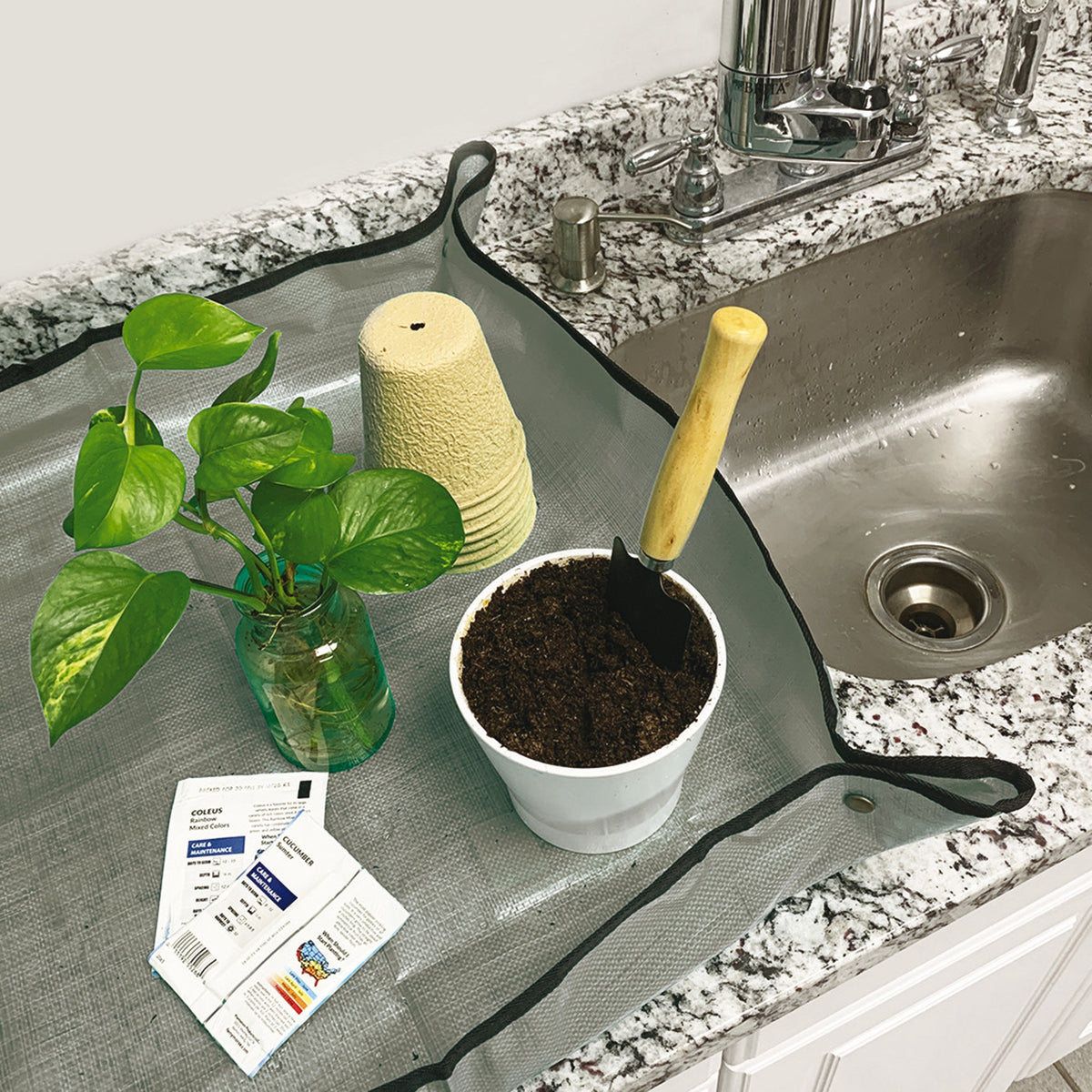 A Tidy Tray on a countertop holding two seed packets, a rooted plant in a green glass jar, a small stack of biodegradable pots, and a white ceramic container with potting soil and a trowel