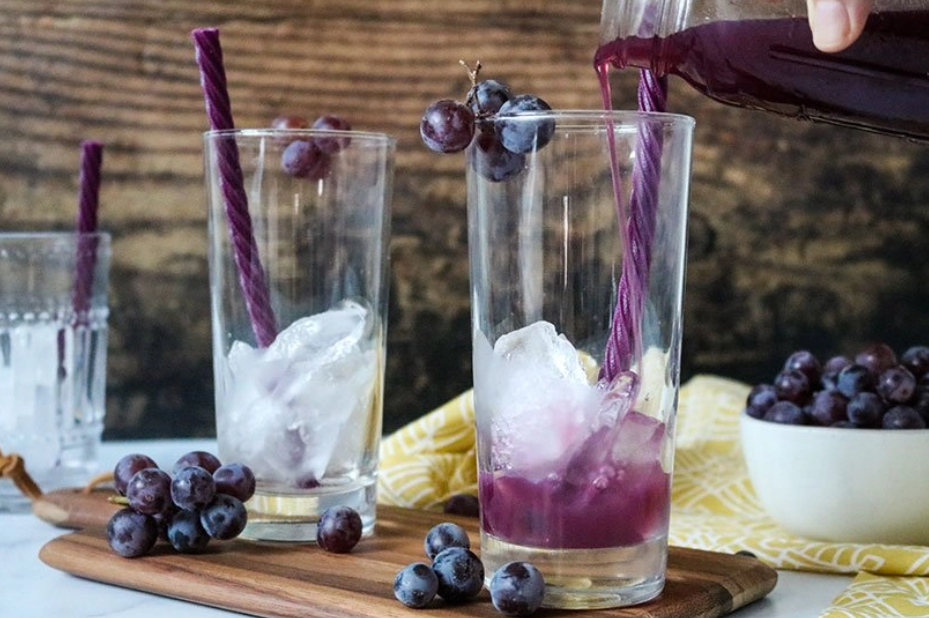 Grape juice being poured into a glass that is holding ice and a Red Vines Grape Licorice Twist