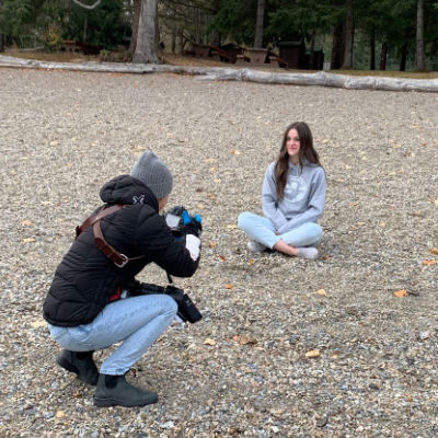 Teenager sitting on the beach in a grey Shuswap Soul Hoodie while a photographer takes a photo