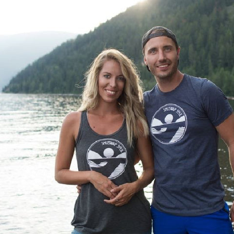 Two people standing and looking at the camera on a beach in Sicamous. The lady is wearing a Shuswap Soul ladies flowy tank in charcoal and the man is wearing a navy unisex tee and a backwards baseball hat