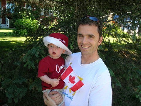 a baby in a red shirt holding a canada flag is in the arms of his father at the South Shuswap Canada Day parade