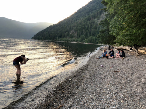 Behind the scenes shot at a photoshoot for Shuswap Soul with Sicamous photographer Moonkissed Memories. The image has the photographer standing in the lake taking photos of a couple sitting on the beach wearing Shuswap Soul hoodies.  