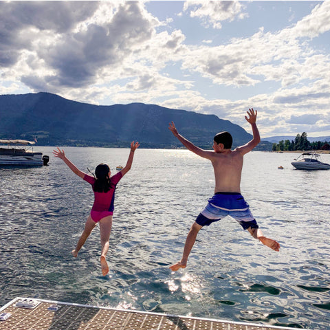 This photo has two kids jumping off a cabin dock with their arms and legs out in a starfish into Shuswap Lake.