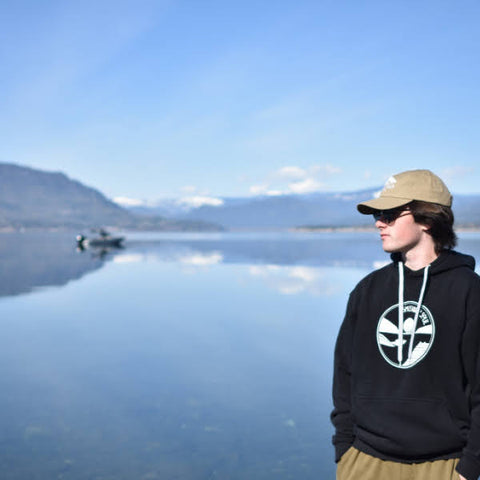 Man standing beside the lake wearing a Shuswap Soul Dad Hat in Khaki and a black Premium Lakeside Hoodie. It's a sunny day, pretty much perfect at the lake. 