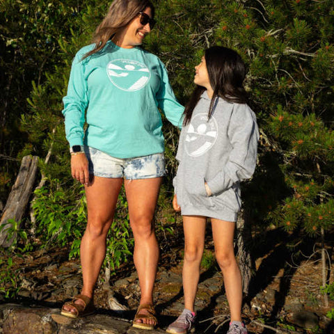 A mom and her daughter are standing at the edge of a forest. The daughter, wearing a Shuswap Soul kids hoodie looks up at her mom who is looking back adoringly at her little girl. 