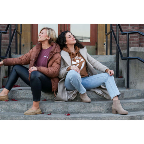 Two women are sitting back to back on the steps of a building in downtown Salmon Arm. Each woman is wearing a Shuswap Soul sweater