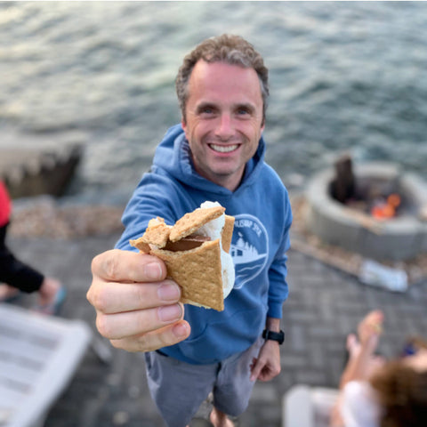 Man holding out a s'more made with marshmallow, chocolate and graham crackers. He's wearing a Shuswap Soul hoodie and is standing by a campfire beside Shuswap Lake