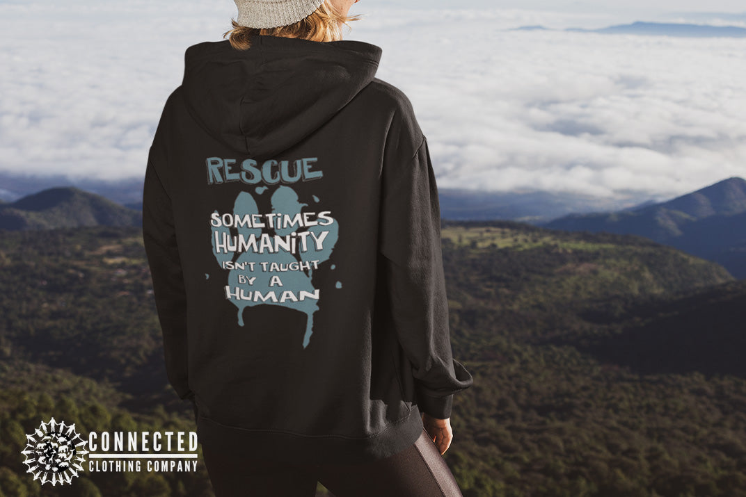 Model wearing Black Show Humanity Unisex Hoodie at the mountains that reads "Rescue. Sometimes humanity isn't taught by a human" - architectconstructor - Ethically and Sustainably Made - 10% donated to the Society for the Prevention of Cruelty to Animals