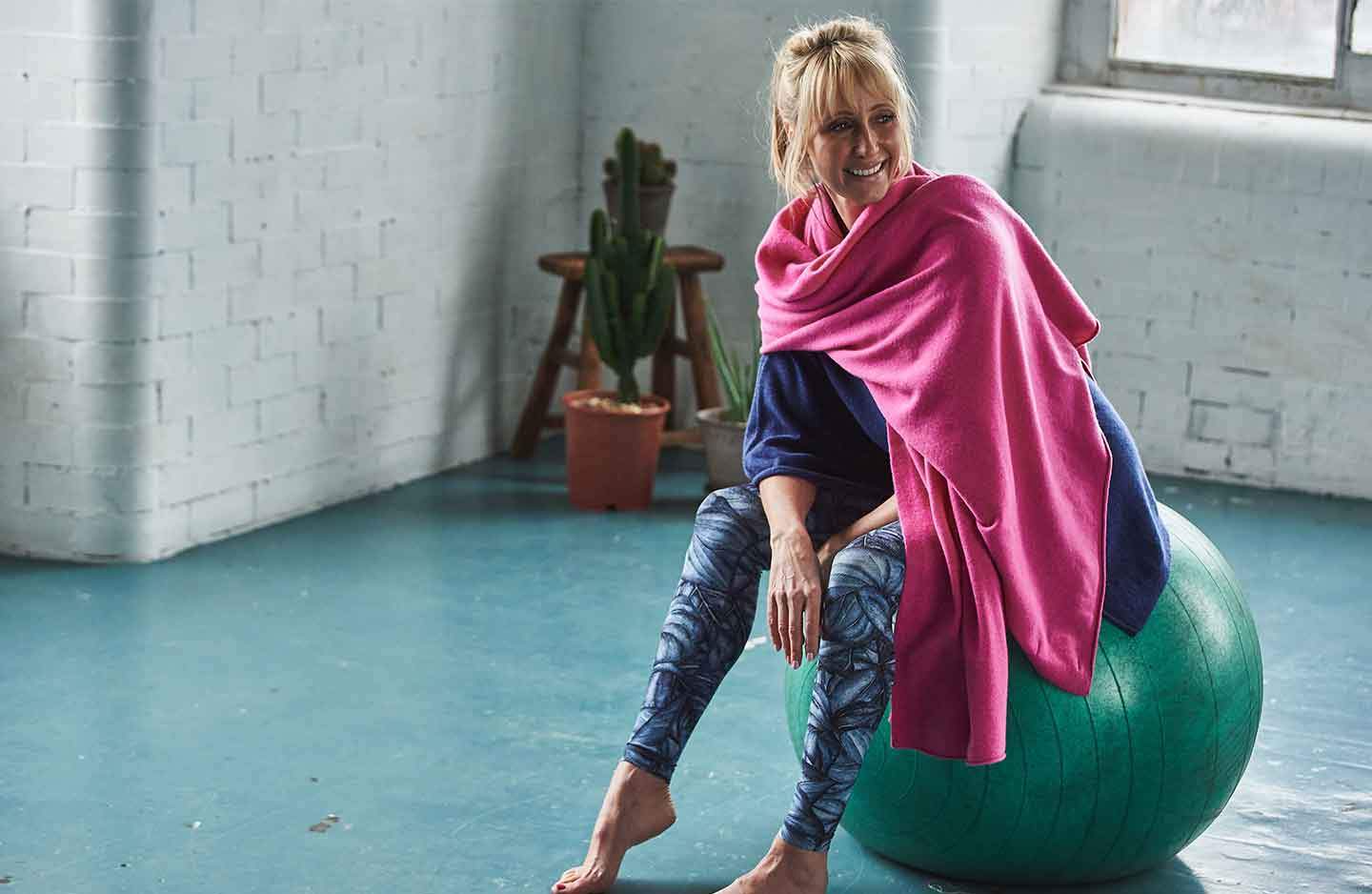 Women sat on green yoga ball in blue sportswear with a blue cashmere poncho layered over the top with a pink cashmere wrap wrapped around shoulders in building with white brick wall and windows with cactus and wooden stool in the corner 