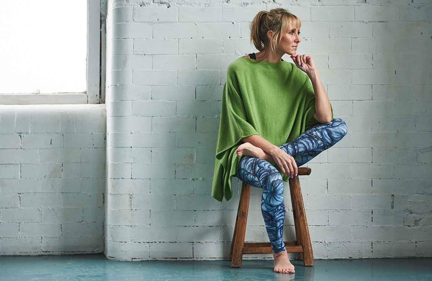 women-sitting-infront-of-a-white-brick-wall-on-a-wooden-stool-wearing-green-cashmere-oversized-poncho-over-sportswear