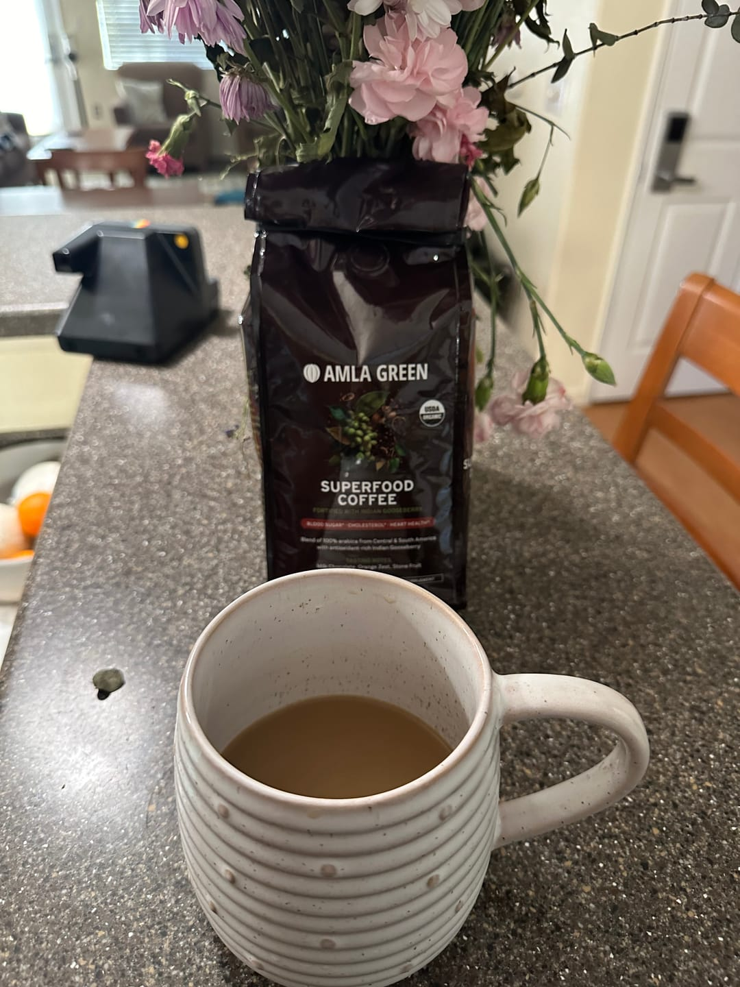 A cup of coffee in a white mug on a counter, near a bag of Amla Green Superfood Coffee.