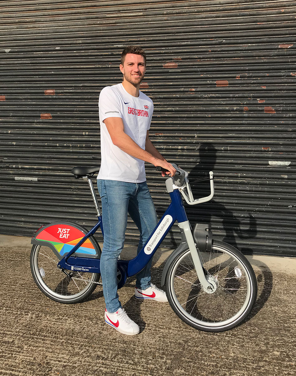 Andrew Pozzi, in a white Great Britain top, astride a just eat branded Transport for London hire bike.
