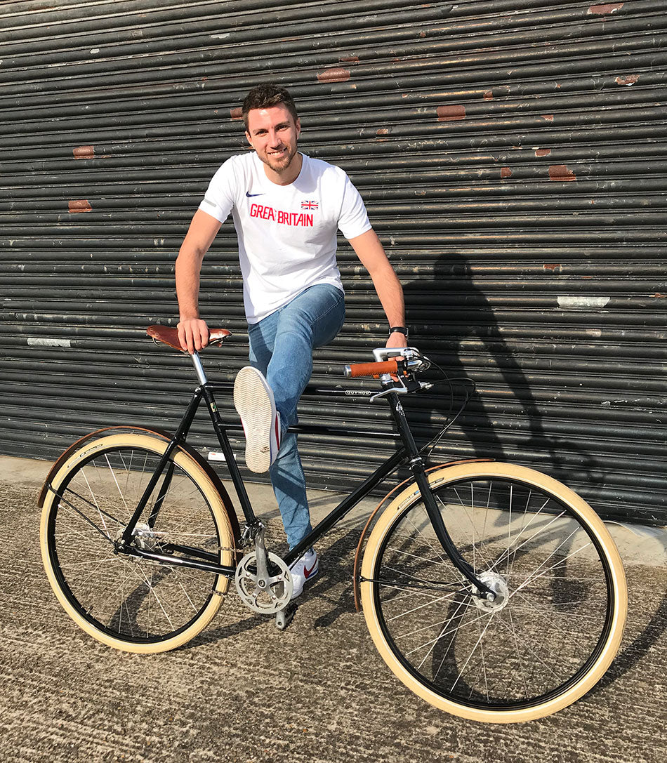 Olympic hurdler, Andrwe Pozzi, pretending to hurdle a double-top tube black Guv'nor bicycle which has wooden mudguards.