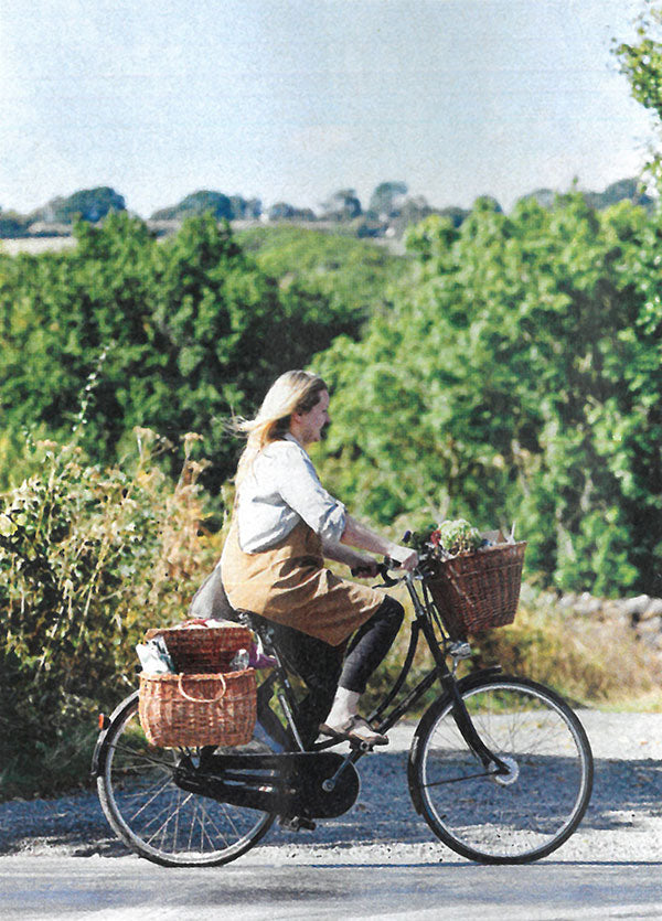 A lady wearing and apron and cycling along a country lane on a classic black bike adorned with wicker baskets.