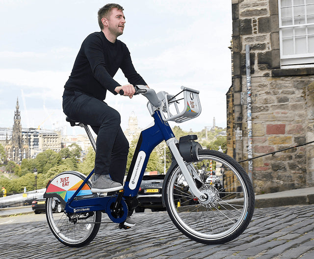A man in black clothing rides a Just Eat bike share e-bike up a cobbled street in Edinburgh.
