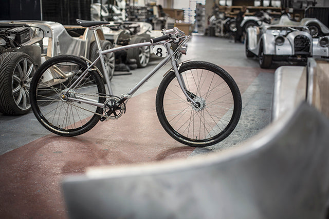 Pashley-Morgan 8 bicycle in silver grey colour next to part-built Morgan motor cars in their factory.