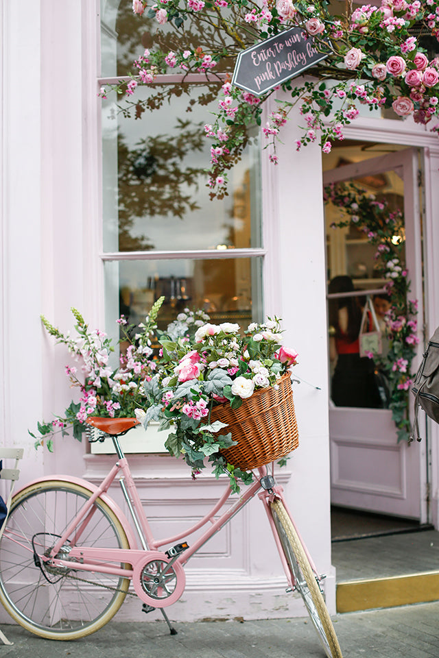 A pink vintage Pashley bicycle with a basket full of pink and white flowers, stood outside a pink cafe shop front.