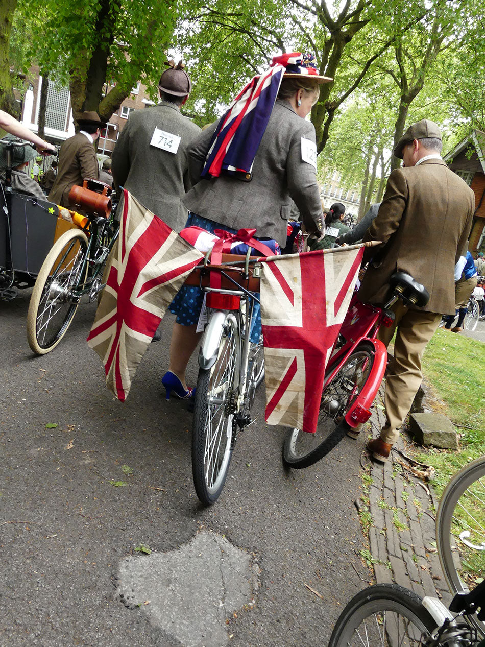 A lady riding her classic bike with two large, vintage union jack flags hanging from her pannier carrier.