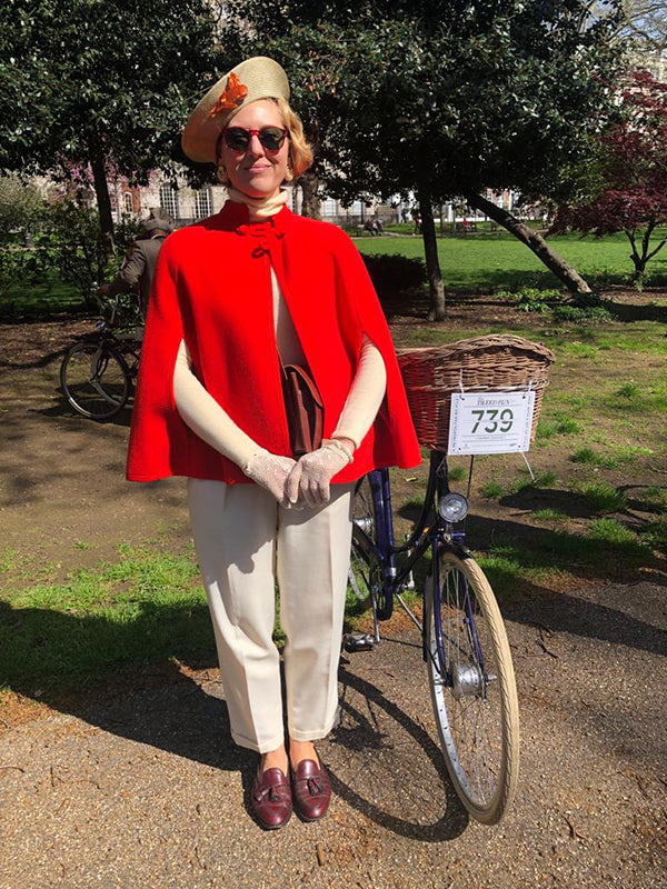 A lady dressed in a vintage wool cape and beret and cream tailored trousers, standing next to her blue Pashley Britannia bike.