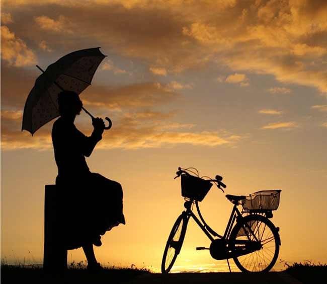 A silhouette of a lady under an umbrella next to her Pashley bicycle with a sunset in the background.