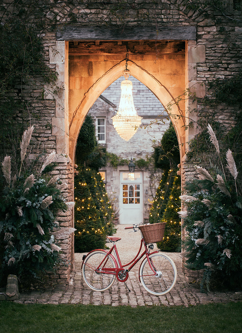 A red Britannia bicycle sat beneath a chandelier hanging from a large stone arch in grand gardens at dusk.
