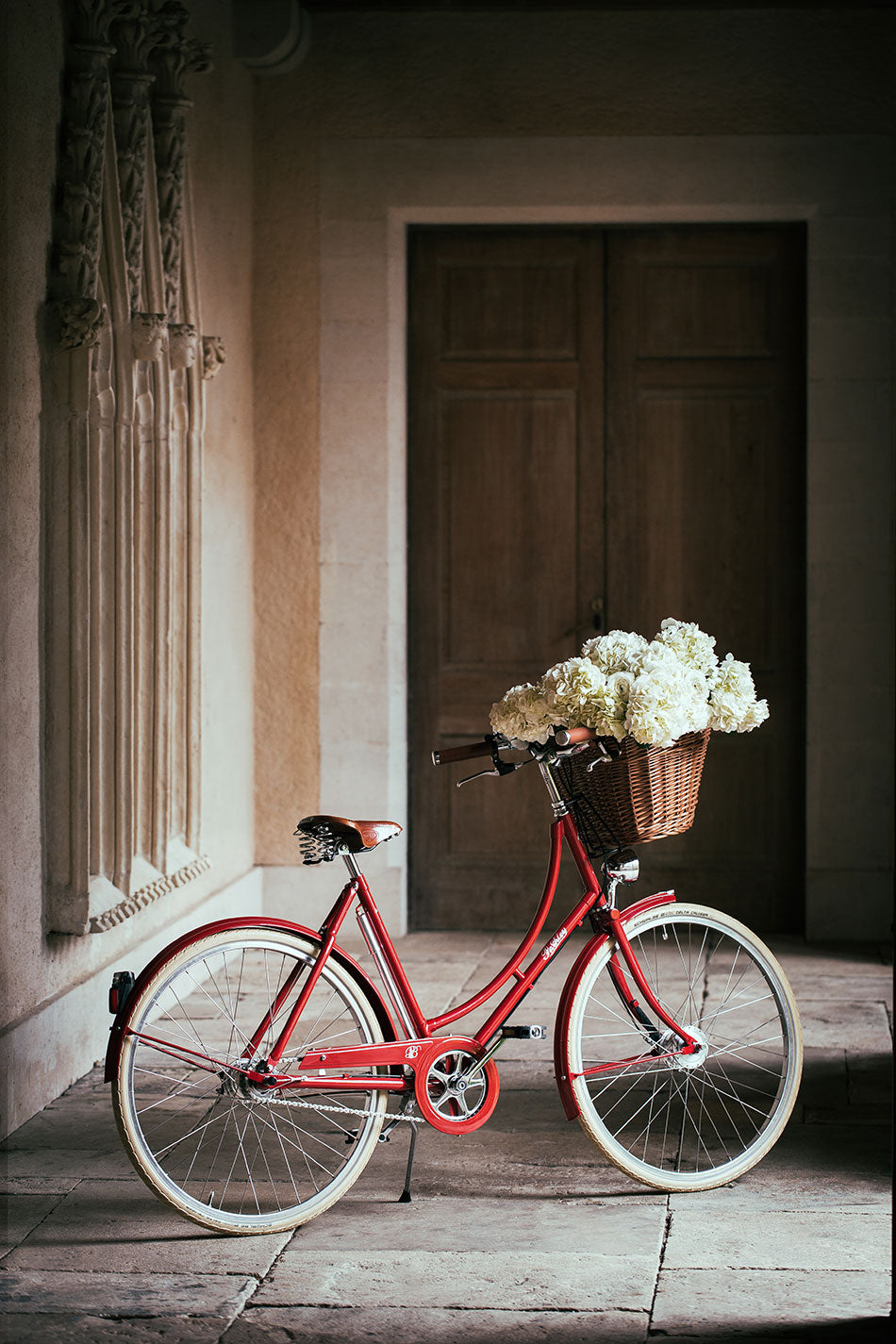 A red vintage bicycle with a leather saddle and large wicker basket full of white hydrangea flowers.