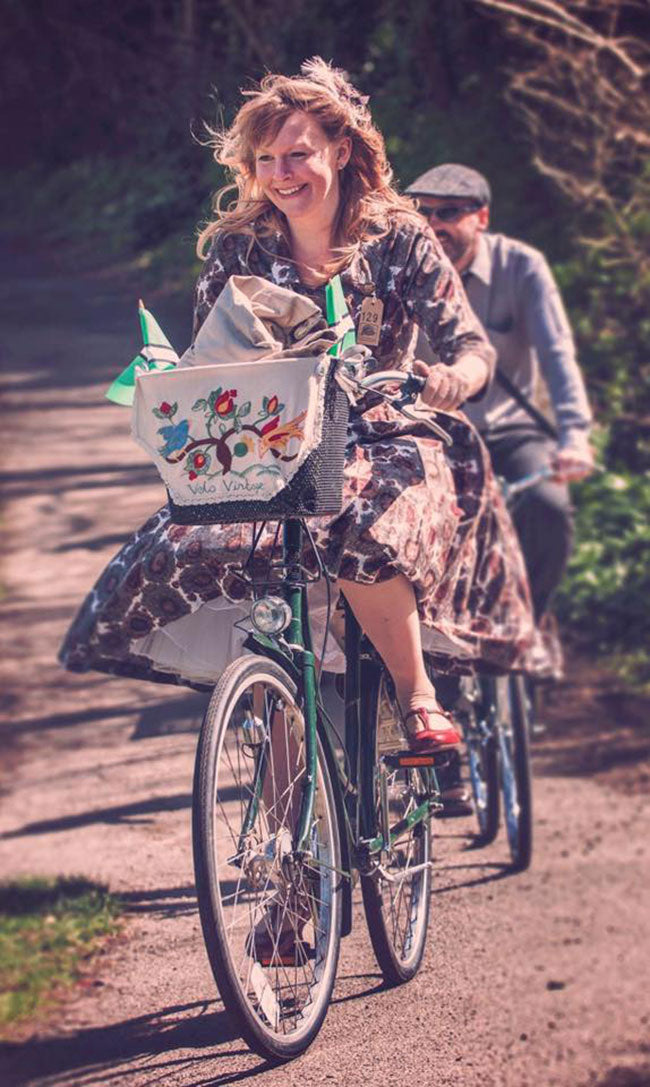 A female cyclist in a vintage floral dress riding her green Pashley princess which she has decorated with an embroidered basket liner.