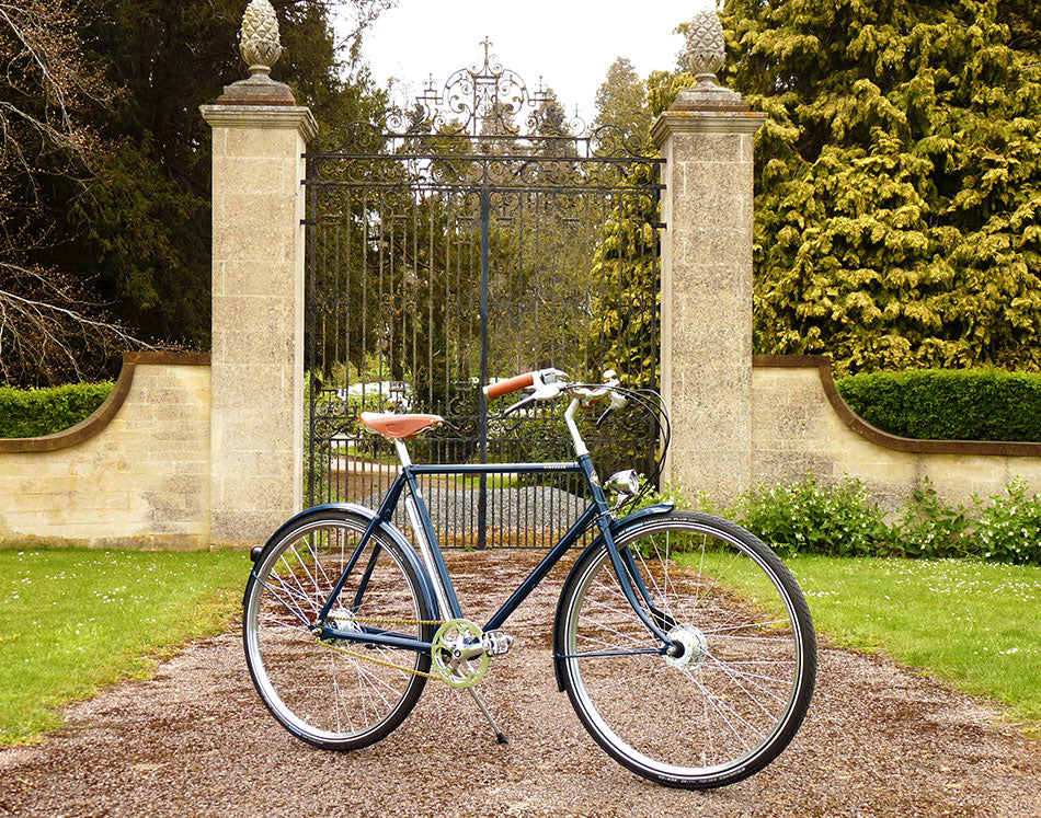 The blue Kingsman bicycle parked at an angle in front of a grand iron gates with stone pillars.