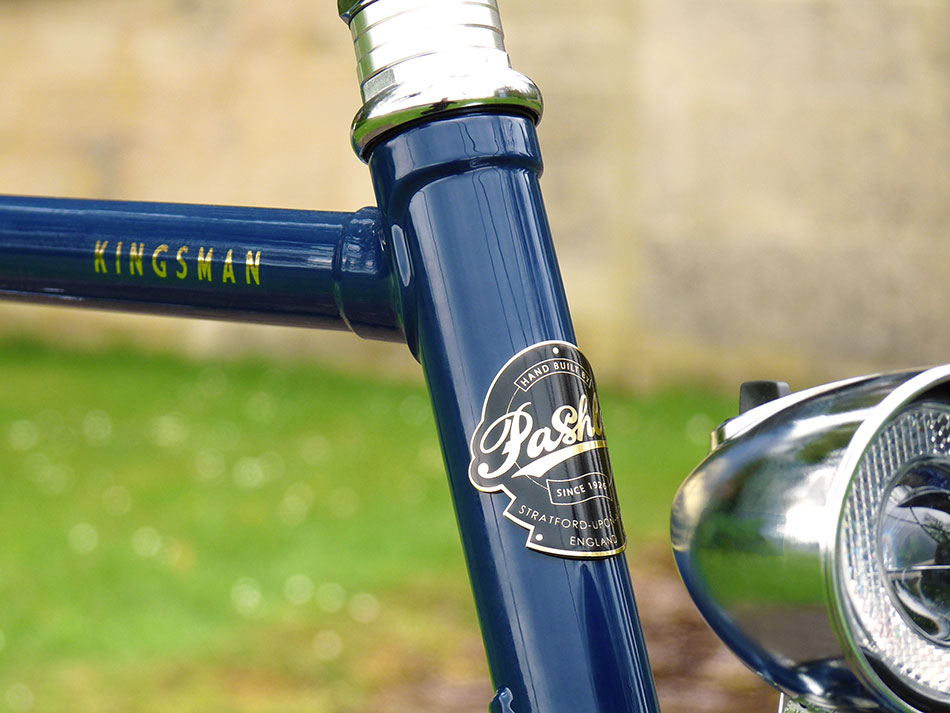 A close-up of the gold Pashley headbadge and Kingsman logo on its Oxford Blue coloured bike frame.