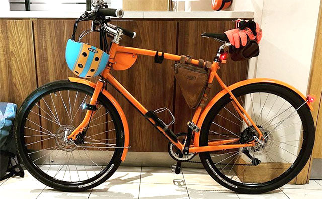 Bright orange bike with black tyres with different bike bags and helmet hanging from handlebars.