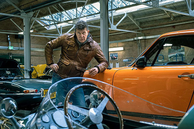 Man in brown leather jacket leaning on a classic orange racing car.