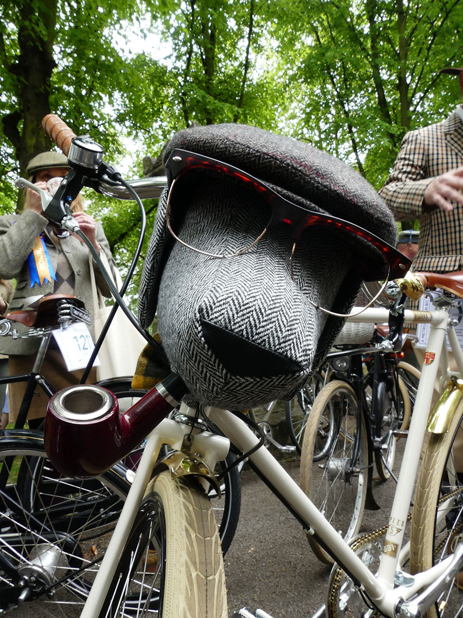 Stuffed dog wearing a flat cap and sunglassse and smoking a pipe, attached to a bicycle's handlebars.