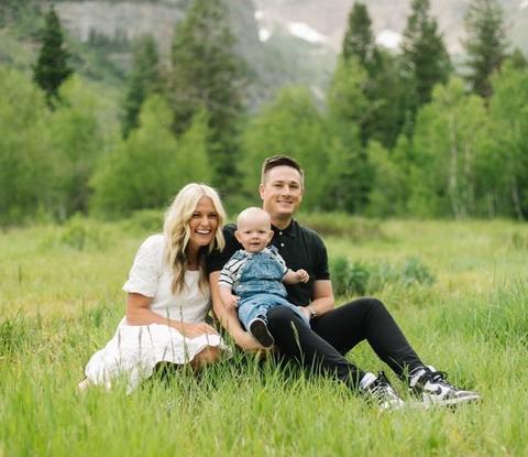 Family photo with mom wearing a white dress, dad in black, and baby wearing overalls in the grass