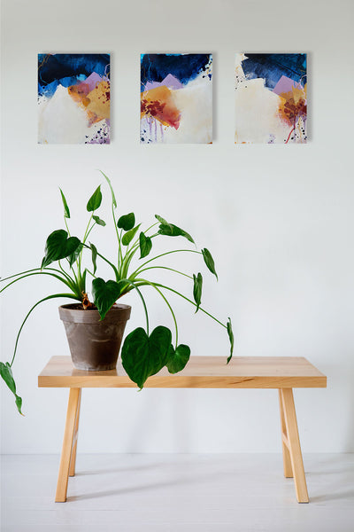 Three coordinating abstract paintings on a white wall above a timber stool with a glossy green monstera pot plant