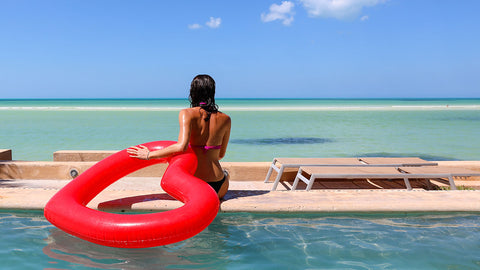 Woman sitting poolside in swimwear holding onto red heart shaped float with her left arm.