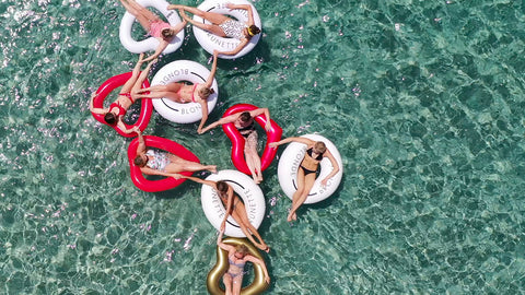 Aerial shot of nine friends in the shallow clear ocean water, floating in red and white floats, all holding hands.