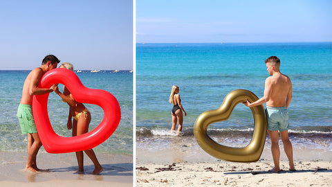 Left: A couple's kiss covered by the red heart float they are holding up while in shallow water. Right: woman gazing back at man holding gold heart float on beach.