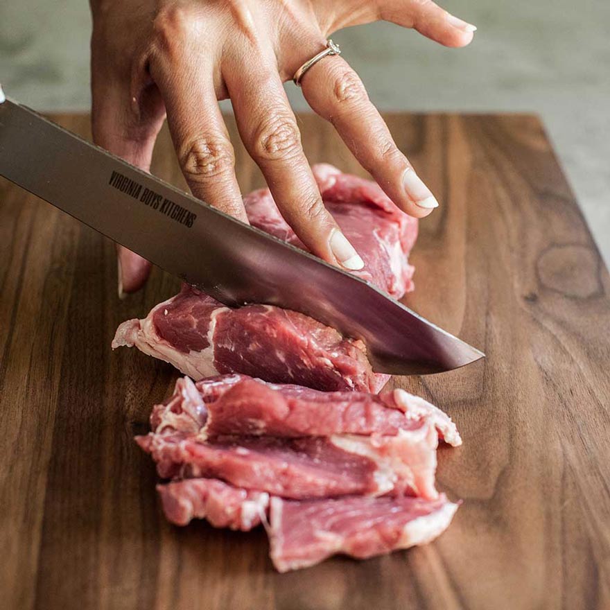 hands using a virginia boys kitchens knife to cut raw steak on a wood chopping board
