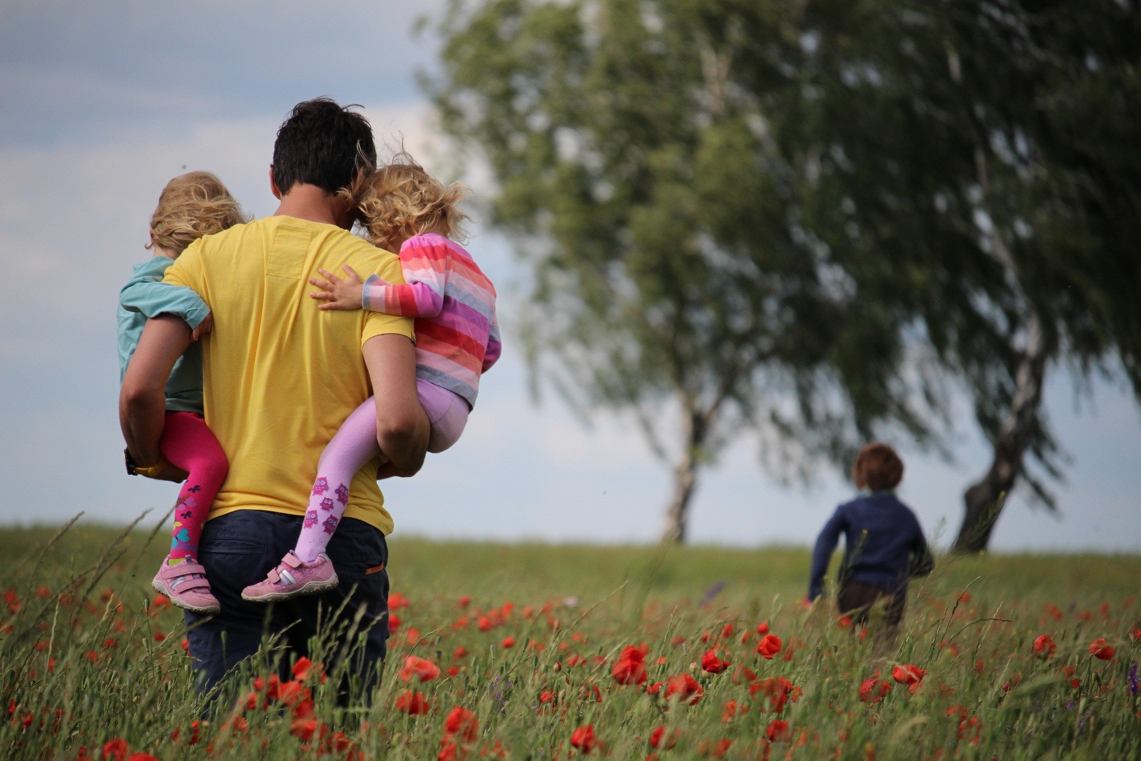 Adult Carrying Two Kids in a Field With Third Kid in The Distance by a Tree