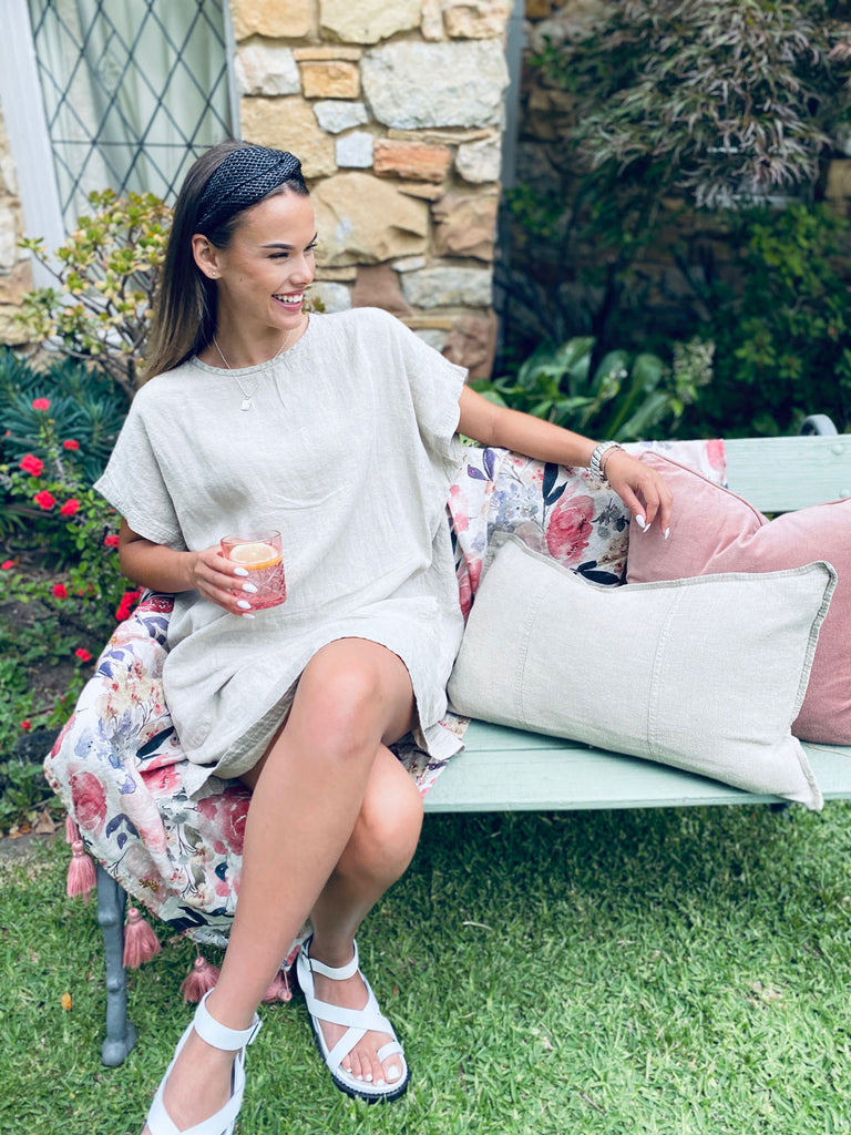 Woman sitting on outdoor seat with cushions and floral throw wearing the Eadie Lifestyle Pocket Linen Dress in natural