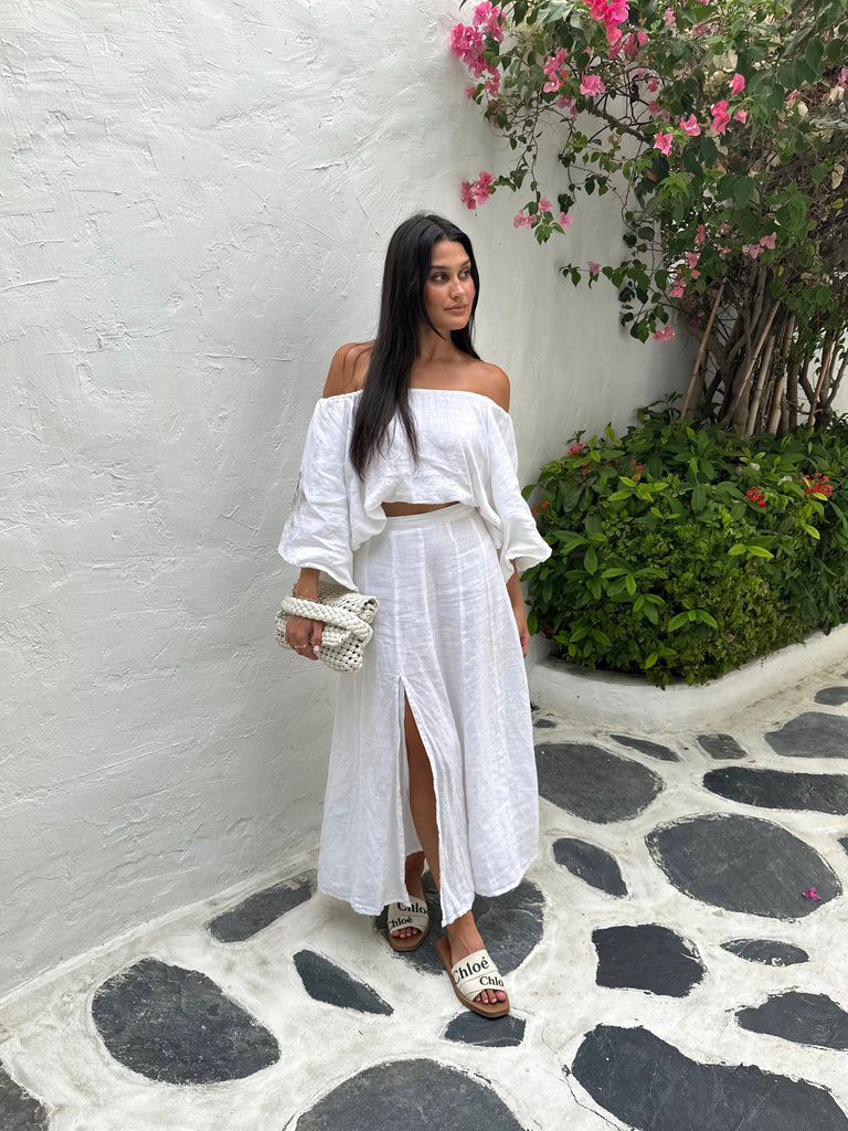 Kate Moore on a cobble stone path in Greece with pink florals in the backdrop. Wearing our white linen Amalfi skirt and matching white linen amalfi top off the shoulder